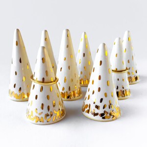 Könnte beinhalten: Sieben weiße Keramik-Ringhalter in Form von Kegeln mit goldenen Punkten und goldenen Akzenten. Die Ringhalter sind in einem Halbkreis auf einer weißen Oberfläche angeordnet.
