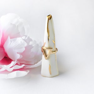 May include: A gold ring with a triangle design sits on a white ceramic ring holder. The holder has a gold leaf design.