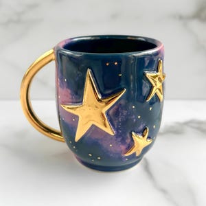 Könnte beinhalten: Eine Keramik-Tasse mit goldenem Henkel und goldenen Sternverzierungen. Die Tasse hat einen dunkelblauen Hintergrund mit einem rosa-violetten Galaxiemuster. Goldene Punkte sind über die Oberfläche verstreut.