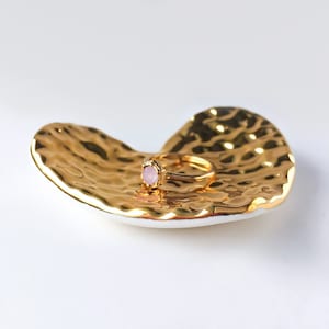 Puede incluir: Un plato dorado en forma de coraz&oacute;n con una superficie texturizada. Un anillo con una piedra rosa descansa sobre el plato.
