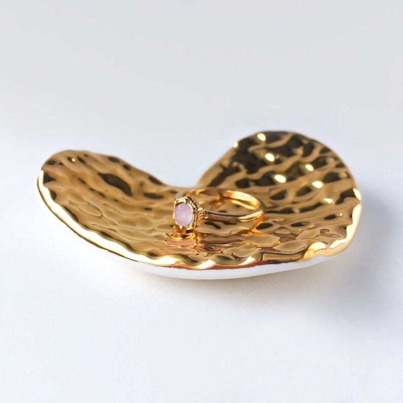 Puede incluir: Un plato dorado en forma de coraz&oacute;n con una superficie texturizada. Un anillo con una piedra rosa descansa sobre el plato.