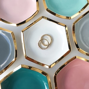 Puede incluir: Una colección de cuencos hexagonales de cerámica en varios colores pastel, cada uno con un borde dorado. Dos anillos dorados descansan en el centro de un cuenco blanco. Los cuencos están dispuestos sobre una superficie blanca.