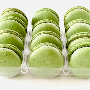 May include: A dozen green macarons in a clear plastic container. The macarons have a light green top and a white bottom. The filling is visible between the two halves of the macaron.