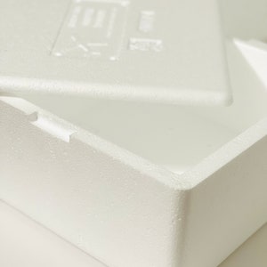 May include: A white, rectangular foam container with a lid slightly ajar. The container has a textured surface and a simple, clean design. The lid features a faint embossed symbol. The container is likely used for shipping or storage.