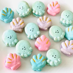 May include: An assortment of colorful macarons in the shape of octopuses and seashells. The macarons are pastel shades of blue, pink, and green, with yellow and black accents. The octopus macarons have smiling faces.