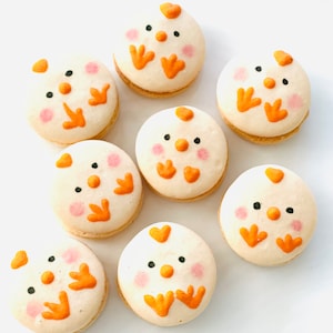 May include: Eight white and orange macarons decorated with chick faces. Each macaron has a different expression and features a small orange beak, black eyes, and orange feet.