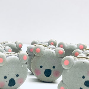May include: A group of gray koala-shaped macarons with pink accents. The macarons have black eyes and noses, and pink cheeks.
