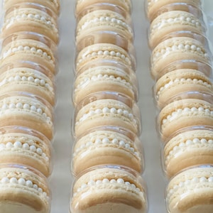 May include: A close-up of a clear plastic container filled with rows of beige macarons decorated with white pearl sprinkles.