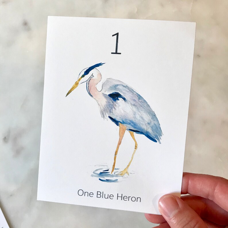 May include: A watercolor print featuring a blue heron standing in water. The bird has blue and gray feathers, a long yellow beak, and long, thin legs. The number "1" is at the top, and the text "One Blue Heron" is at the bottom.