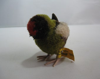 Könnte beinhalten: Ein kleiner grüner, brauner und weißer Stoffvogel mit rotem Kopf. Der Vogel hat ein gelbes Etikett mit der Aufschrift "The Original Steiff 712210".