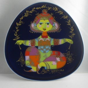 May include: A blue and gold ceramic plate featuring a colourful illustration of a woman in traditional dress. The plate is decorated with gold leaf and has a floral design around the edge. The artist's signature, "Bjorn Wiinblad", is visible in the bottom right corner.