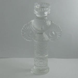 May include: A clear glass candle holder with a decorative design. The holder is shaped like a person with a wide base and a narrow top.