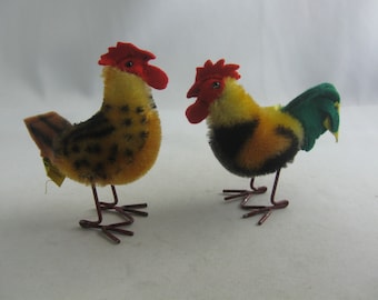 May include: Two plush toy roosters with wire legs. One rooster is yellow, black, and green. The other rooster is yellow and black.