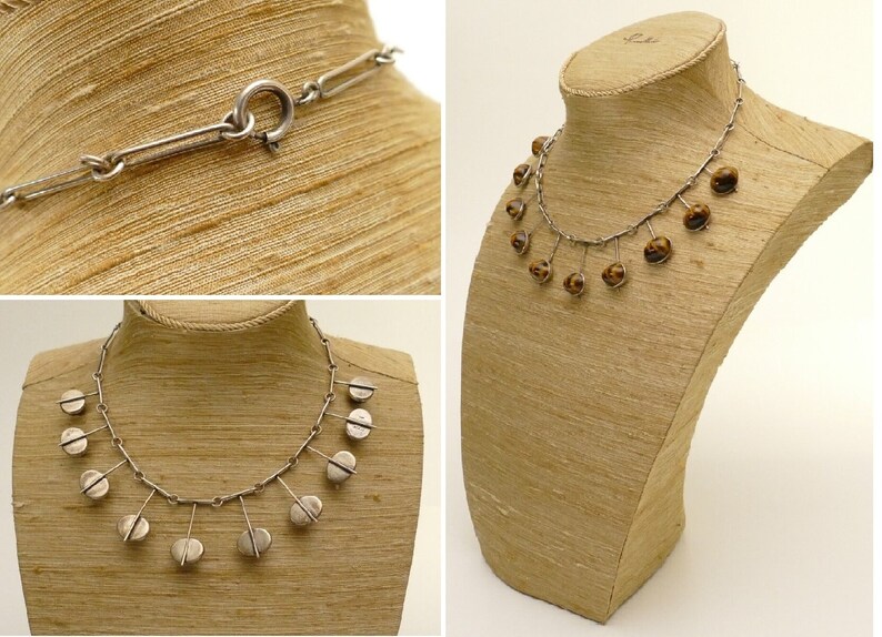 May include: A silver necklace with a chain and dangling round silver charms. The necklace is displayed on a brown mannequin.