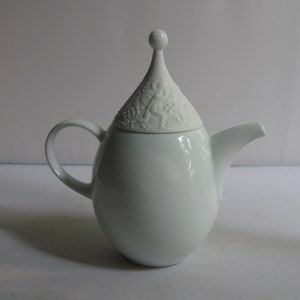 May include: White porcelain teapot with a decorative, ornate lid. The lid has a pointed top and a raised design.