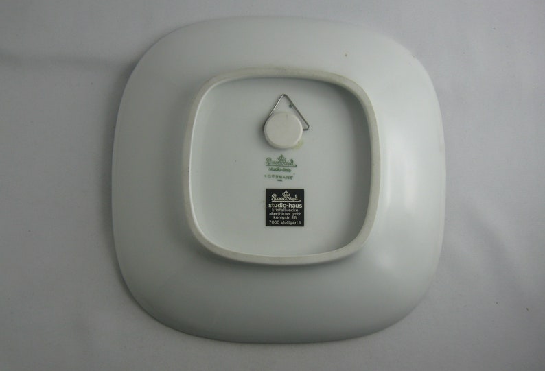 May include: White square ceramic plate with a green and black label that reads "studio-haus, keramik-werke, k&ouml;nigstr. 49, 7000 stuttgart 1".