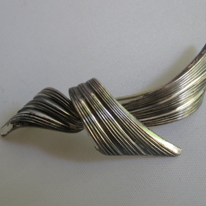 May include: A silver brooch with a twisted, ribbon-like design. The brooch is made of textured metal and has a polished finish.