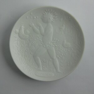 May include: White ceramic plate with a raised design of a figure with a peacock and other birds. The plate is inscribed with the word "Papgeno" at the bottom.