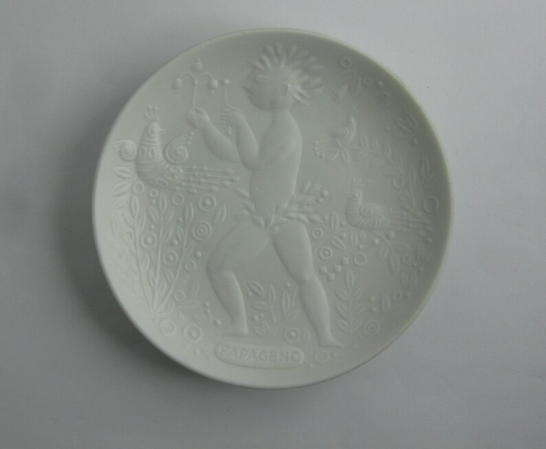 May include: White ceramic plate with a raised design of a figure with a peacock and other birds. The plate is inscribed with the word "Papgeno" at the bottom.