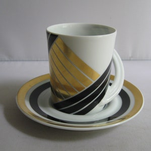 Puede incluir: Una taza de espresso y un platillo de porcelana blanca con un diseño geométrico en negro y dorado. La taza tiene un asa única que se extiende desde el lado.