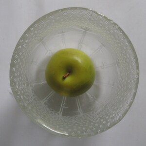 Op de afbeelding: Een groene appel in een glazen kom met een getextureerd oppervlak. De kom heeft een decoratief patroon van kleine cirkels.