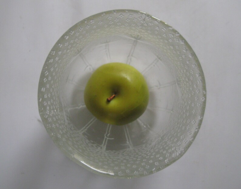 Op de afbeelding: Een groene appel in een glazen kom met een getextureerd oppervlak. De kom heeft een decoratief patroon van kleine cirkels.