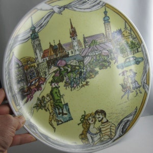 May include: A decorative plate with a yellow background featuring a detailed illustration of a European town scene. The plate includes images of buildings, people, and a market. The rim is white with a gray border. The plate is held by a hand.