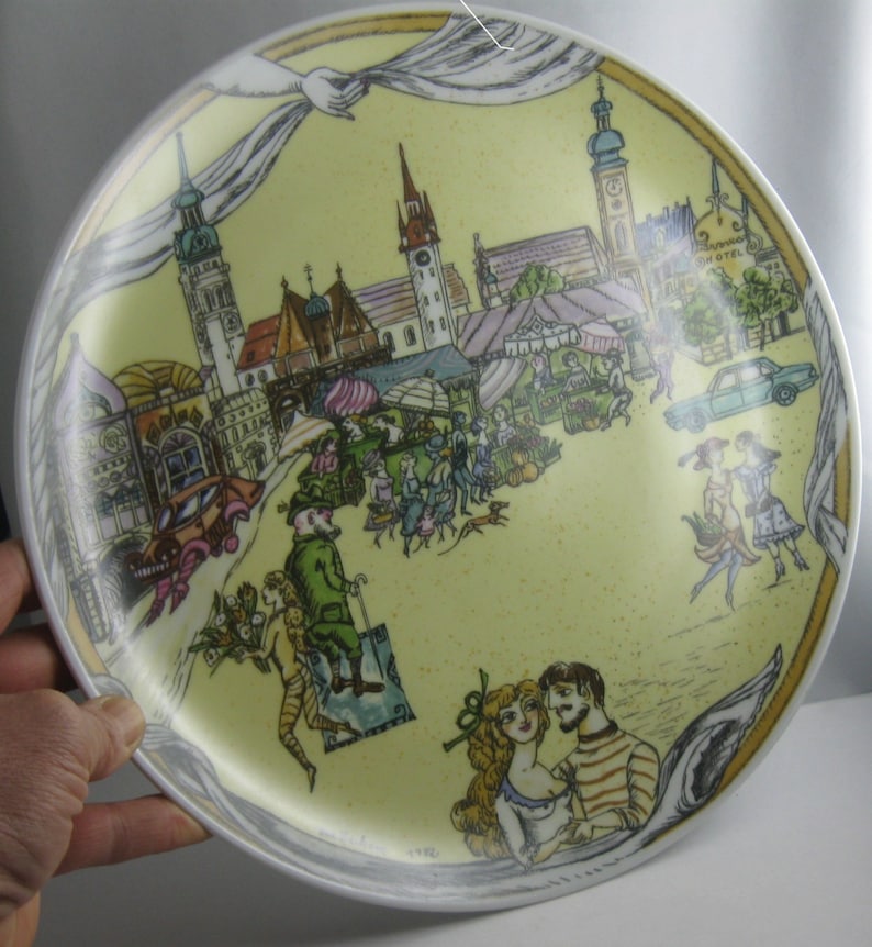 May include: A decorative plate with a yellow background featuring a detailed illustration of a European town scene. The plate includes images of buildings, people, and a market. The rim is white with a gray border. The plate is held by a hand.