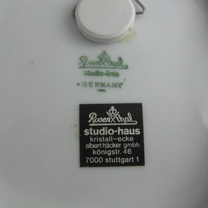 May include: Close-up of a white porcelain plate with two green and black labels. The top label reads "Rosenthal studio-linie GERMANY". The bottom label reads "Rosenthal studio-haus kristall-ecke albert h&auml;cker gmbh k&ouml;nigstr. 46 7000 stuttgart 1".