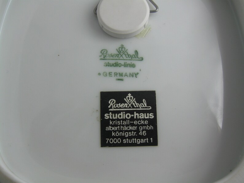 May include: Close-up of a white porcelain plate with two green and black labels. The top label reads "Rosenthal studio-linie GERMANY". The bottom label reads "Rosenthal studio-haus kristall-ecke albert h&auml;cker gmbh k&ouml;nigstr. 46 7000 stuttgart 1".