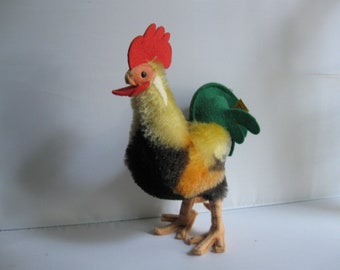 May include: A plush toy rooster with a red comb, yellow and black feathers, and a green felt tail. The rooster has a small tag on its wing.