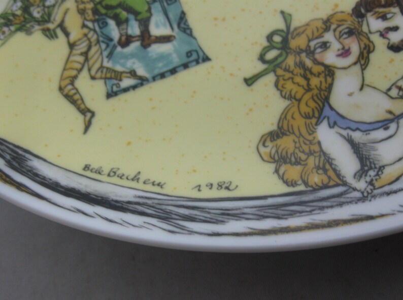 May include: A decorative plate with a yellow background and a detailed illustration. The artwork features figures in a classical style, with a woman with blonde hair and a man with a beard. The plate is signed "Belle Bachere" and dated "1982".