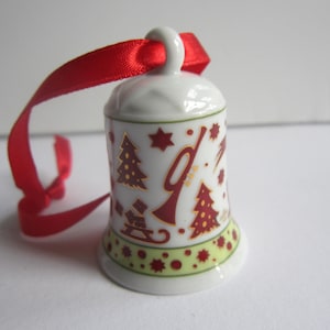 Op de afbeelding: Een witte keramische bel-ornament met rode en gouden kerstontwerpen, waaronder bomen en sterren. Een rood lint is bevestigd om op te hangen. De bel heeft een groen en gele rand.
