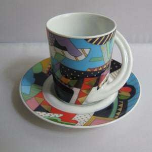May include: A colorful ceramic coffee cup and saucer set. The cup and saucer feature a geometric design with blue, purple, red, and gold accents. The cup has a unique handle design.