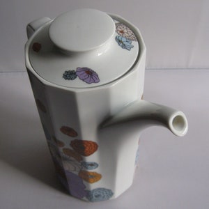 May include: White ceramic teapot with a geometric design and a lid. The teapot is decorated with colorful floral patterns in orange, purple, and blue. The spout is curved, and the handle is not visible.