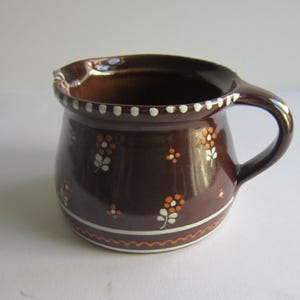 Puede incluir: Una jarra de cerámica marrón con un asa blanca y diseños florales blancos y naranjas.