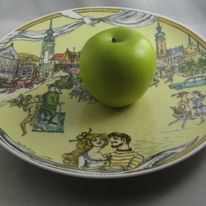May include: A decorative ceramic plate with a yellow background and a detailed illustration of a European town scene. A green apple rests on the plate. The plate's design includes figures, buildings, and vehicles.