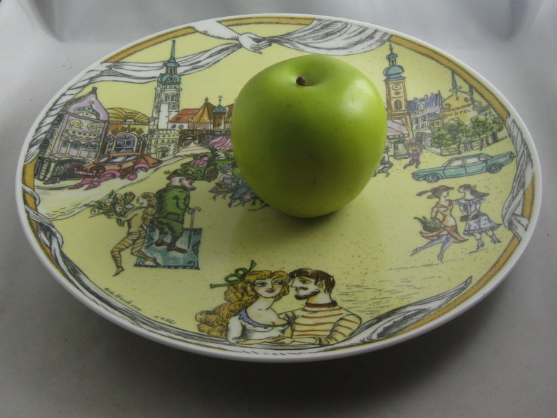 May include: A decorative ceramic plate with a yellow background and a detailed illustration of a European town scene. A green apple rests on the plate. The plate's design includes figures, buildings, and vehicles.