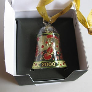 Op de afbeelding: Een glazen kerstversiering in de vorm van een bel met een gouden lint. De bel is versierd met een zwarte en rode viool, gouden sterren en het jaar 2000.