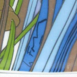 May include: Close-up of a blue and brown patterned ceramic plate with a green leaf design. The plate has a white rim and a signature in the lower right corner.