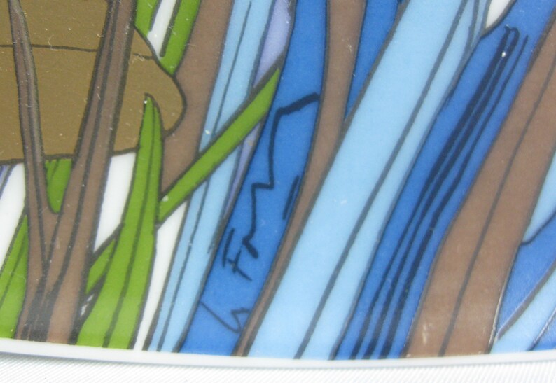 May include: Close-up of a blue and brown patterned ceramic plate with a green leaf design. The plate has a white rim and a signature in the lower right corner.