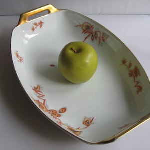 Könnte beinhalten: Eine weiße Porzellanservierplatte mit goldfarbenem Rand und Blumenmuster. Die Platte hat zwei Griffe und ist mit orangefarbenen, roten und grünen Blumendesigns verziert. Ein grüner Apfel liegt in der Mitte der Platte.