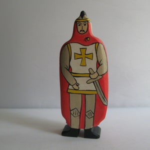 Könnte beinhalten: Eine hölzerne Spielzeug-Ritterfigur mit einem roten Umhang, einer weißen Rüstung und einem gelben Kreuz. Der Ritter hält ein Schwert und trägt einen schwarzen Helm mit einer spitzen Spitze.