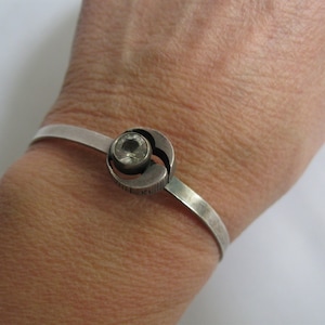 Puede incluir: Un brazalete de plata con un cristal transparente engastado en un círculo de plata. El círculo está unido al brazalete con una bisagra de plata.