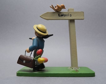 Op de afbeelding: Een houten figuur van een persoon met een strohoed en een bruine koffer. Het figuur staat naast een houten wegwijzer met het woord "Leipzig" erop. Een kleine vogel zit bovenop de wegwijzer.