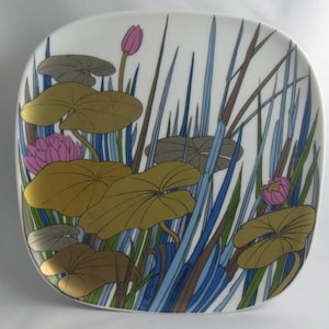 May include: A white ceramic plate with a stylized design of pink water lilies, green and brown lily pads, and blue and brown reeds.