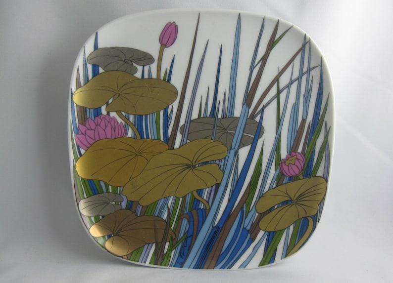 May include: A white ceramic plate with a stylized design of pink water lilies, green and brown lily pads, and blue and brown reeds.