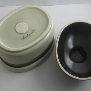 May include: A set of two ceramic bowls, one white with a brown rim and the other black with a white rim. The white bowl has a gold sticker that reads "Handgemalt".