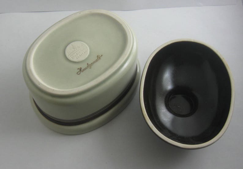 May include: A set of two ceramic bowls, one white with a brown rim and the other black with a white rim. The white bowl has a gold sticker that reads "Handgemalt".