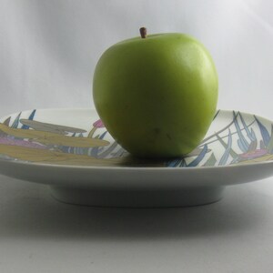 May include: A white ceramic serving dish with a green apple on top. The dish has a floral design with blue, green, yellow, and pink accents.